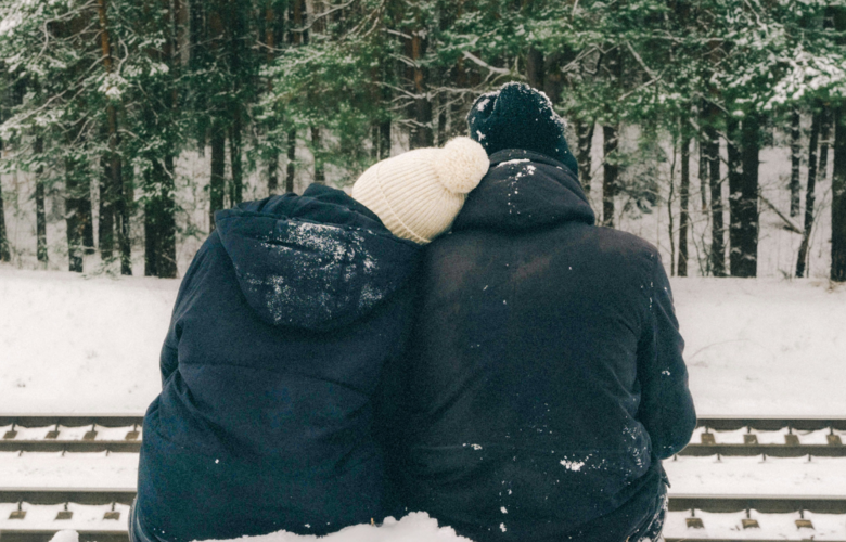 Ein Pärchen kuschelt sich aneinander. Sie sitzen im Schnee und blicken auf den verschneiten Wald.