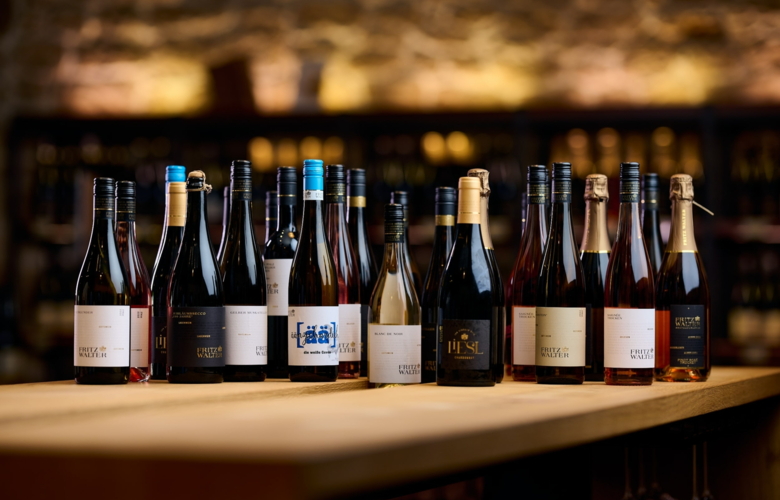 Mehrere Weinflaschen unterschiedlicher Formen und Farben stehen nebeneinander auf einem Holztisch vor einer unscharfen Steinwand.