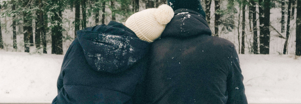 Ein Pärchen kuschelt sich aneinander. Sie sitzen im Schnee und blicken auf den verschneiten Wald.