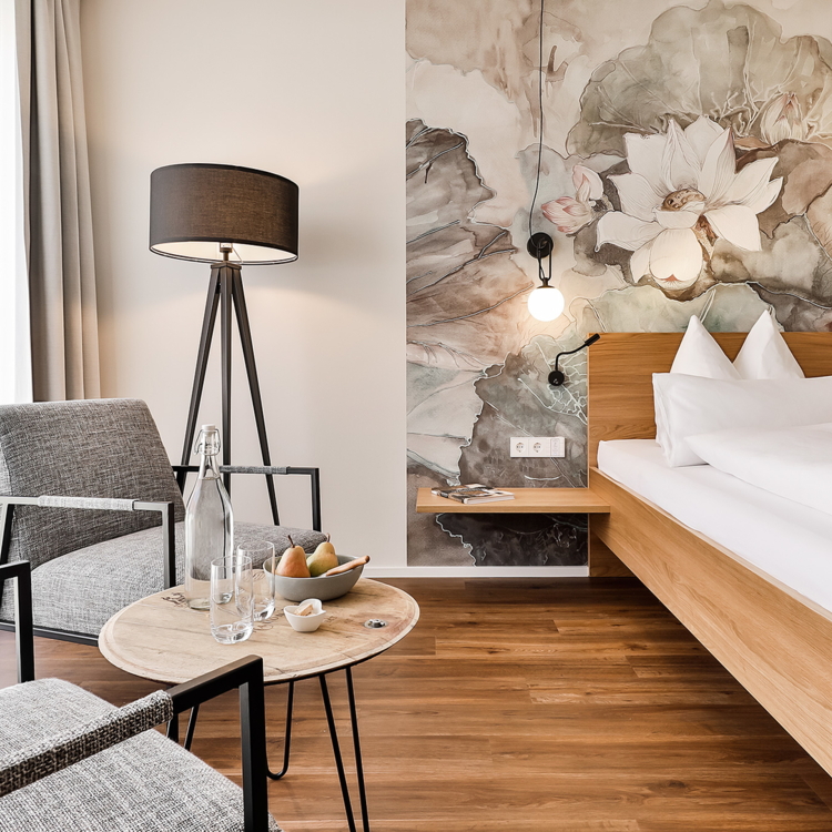 Ein modernes Hotelzimmer mit sanften Farben und einem schönen Blumenmuster an der Wand. Es gibt ein komfortables Bett und einen gemütlichen Sitzbereich mit Stühlen.