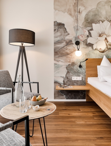 Ein modernes Hotelzimmer mit sanften Farben und einem schönen Blumenmuster an der Wand. Es gibt ein komfortables Bett und einen gemütlichen Sitzbereich mit Stühlen.