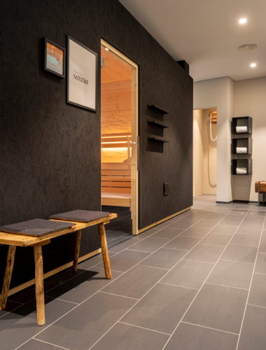 Ein moderner Flur mit angenehmem Ambiente, der zur Sauna im Wellnessbereich des WeinHotels FRITZ WALTER führt. Die Wände sind dunkel, und es gibt ein paar praktische Möbelstücke sowie Dekorationselemente.