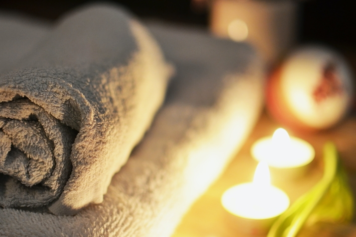 Zwei aufgerollte Handtücher liegen neben brennenden Teelichtern und einer lila Tulpe auf einem Holztisch, unscharfer Hintergrund.