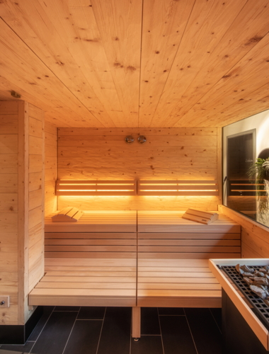 Ein modernes Sauna-Ambiente mit warmem Holz und sanftem Licht. Die große Fensterfront bietet einen Blick nach draußen und schafft eine entspannende Atmosphäre.