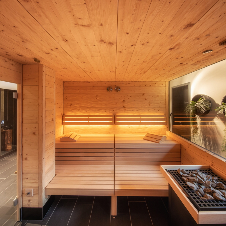 Ein modernes Sauna-Ambiente mit warmem Holz und sanftem Licht. Die große Fensterfront bietet einen Blick nach draußen und schafft eine entspannende Atmosphäre.