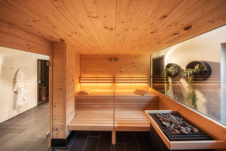 Ein modernes Sauna-Ambiente mit warmem Holz und sanftem Licht. Die große Fensterfront bietet einen Blick nach draußen und schafft eine entspannende Atmosphäre.