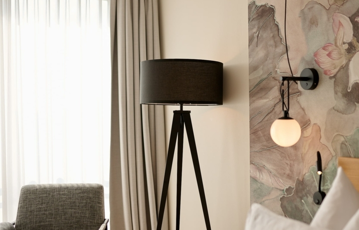 Ein Bett mit weißer Bettwäsche im Vordergrund. Im Hintergrund gibt es eine Sitzgelegenheit und eine Stehlampe.