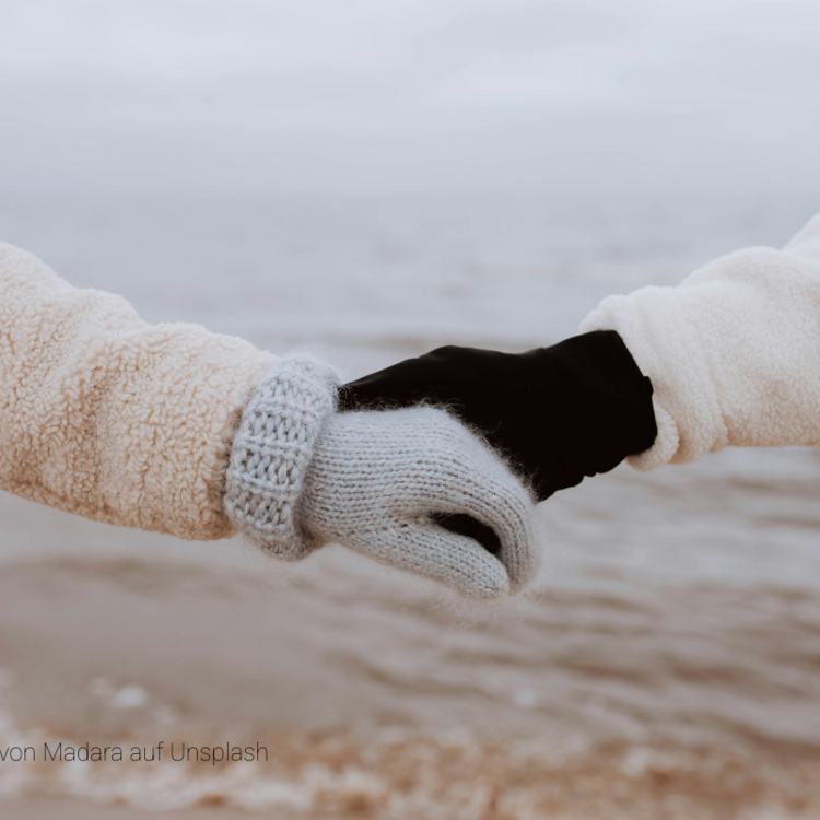 Zwei Hände in Winterkleidung halten sich vor einem unscharfen Gewässer fest, eine Hand trägt grauen Strickhandschuh, die andere schwarzen Handschuh.