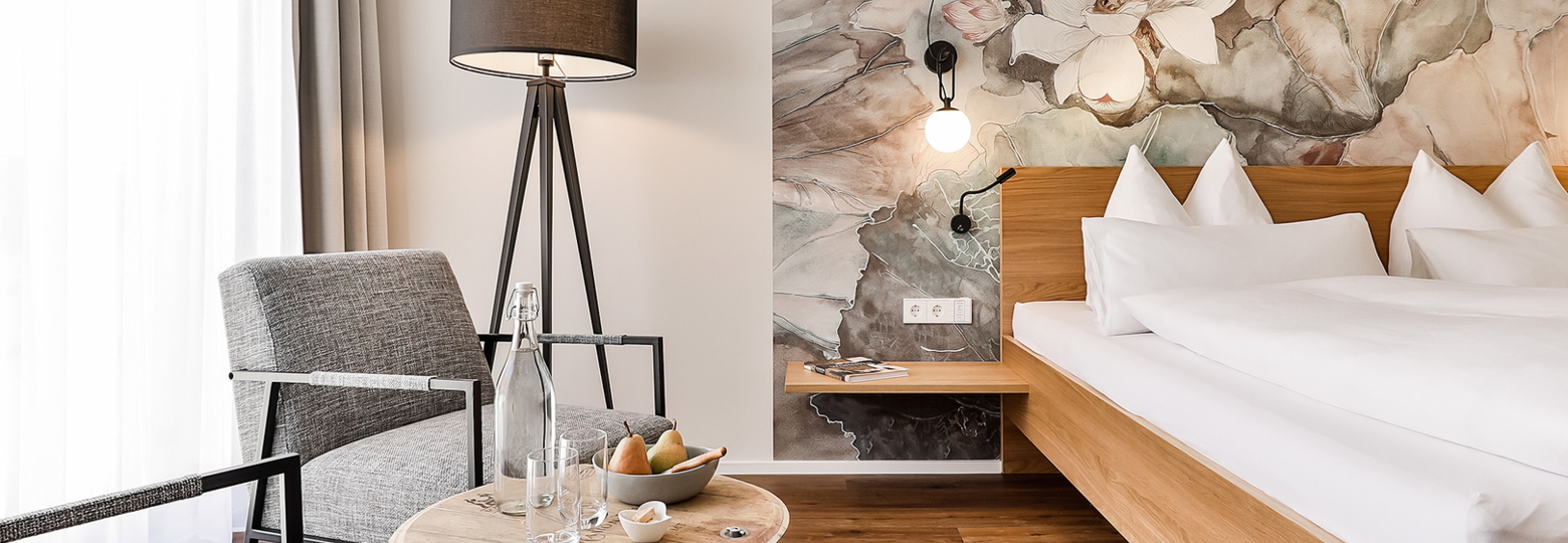 Ein modernes Hotelzimmer mit sanften Farben und einem schönen Blumenmuster an der Wand. Es gibt ein komfortables Bett und einen gemütlichen Sitzbereich mit Stühlen.