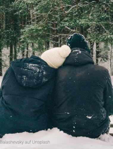 Ein Pärchen kuschelt sich aneinander. Sie sitzen im Schnee und blicken auf den verschneiten Wald.