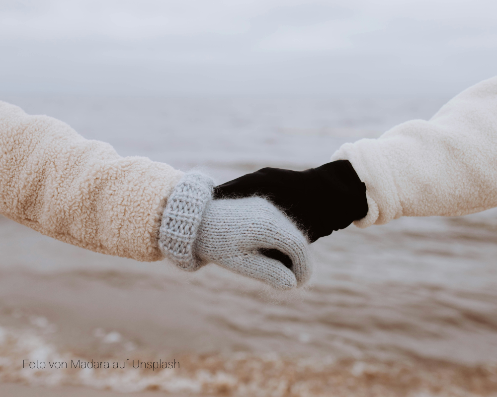 Zwei Hände in Winterkleidung halten sich vor einem unscharfen Gewässer fest, eine Hand trägt grauen Strickhandschuh, die andere schwarzen Handschuh.